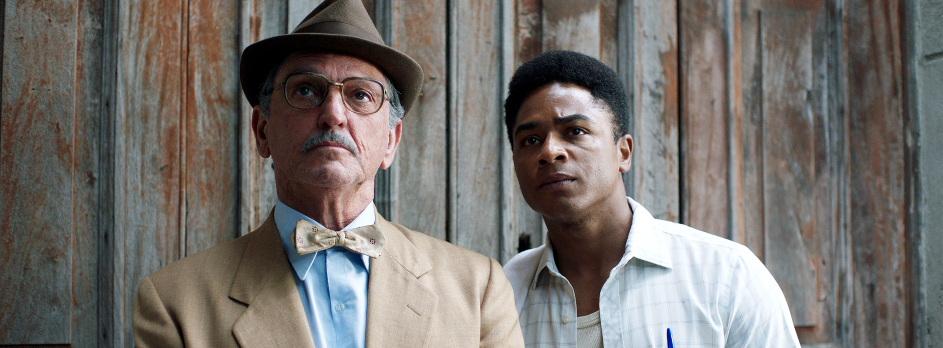 Em cena do filme Saudosa Maloca, o ator Paulo Miklos aparece caracterizado como Adoniran Barbosa, de bigode, terno bege, camisa azul e uma gravata borboleta bege, além de óculos e chapéu. Ele está ao lado do ator Sidney Santiago, que é negro, e usa uma ca