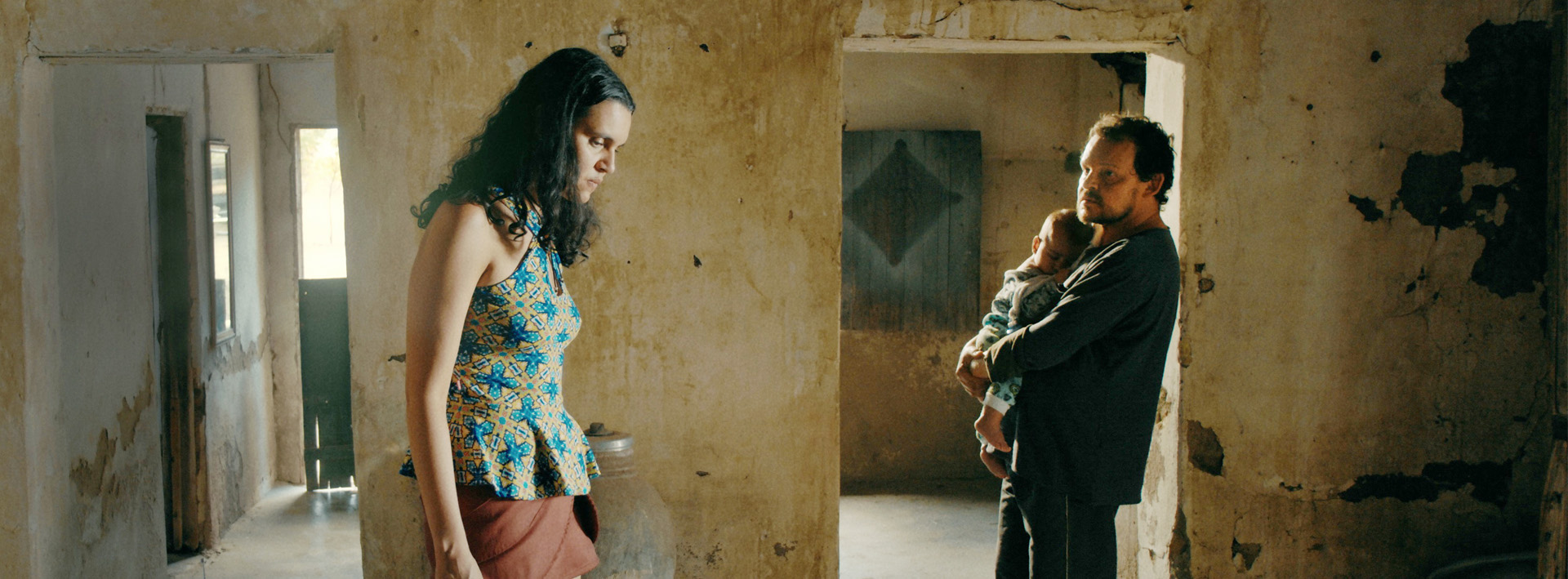 Em cena do filme Mais Pesado É o Céu, uma mulher e um homem, ambos brancos e de cabelo pretos estão dentro de uma casa abandonada. A está olhando para o chão, enquanto o homem, mais ao fundo da imagem, olha para a mulher e segura um bebê no colo.