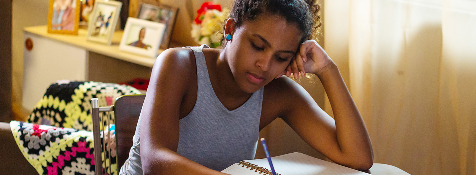 Em cena do filme Eu Sou Maria, uma garota jovem negra de regata cinza está sentada, com a cabeça apoiada na mão esquerda, enquanto escreve a lápis em um caderno.