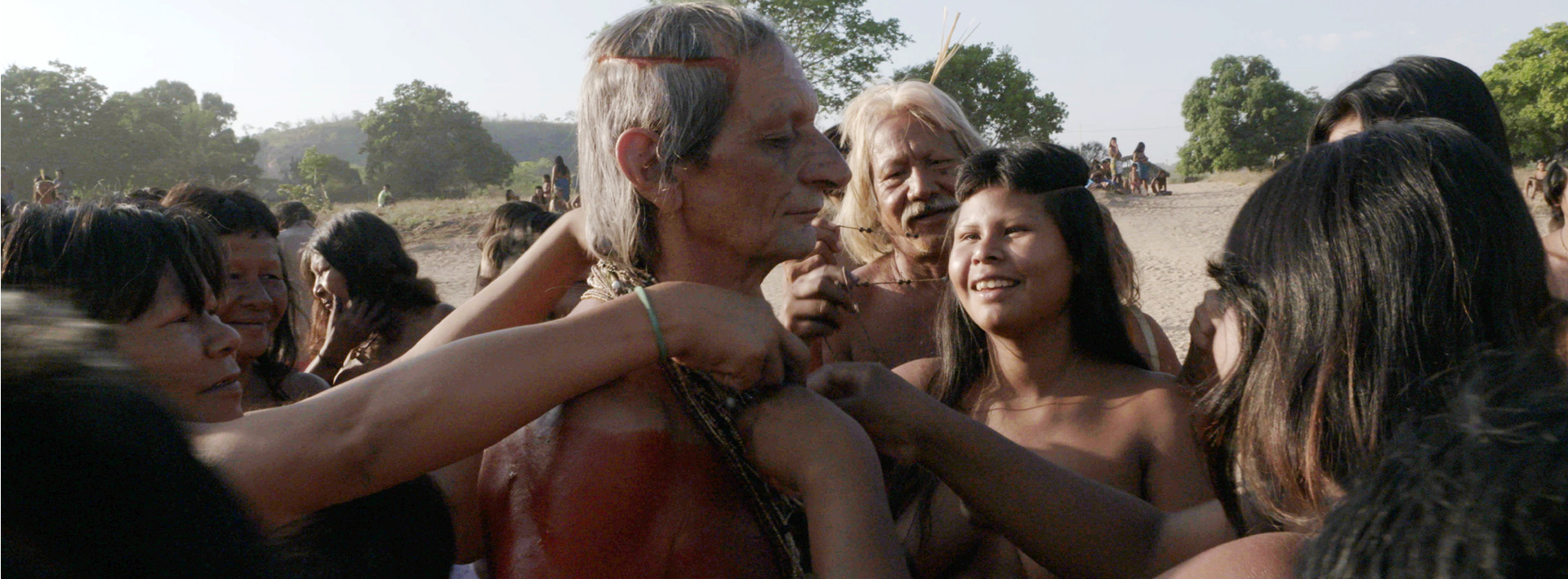 Em cena do filme De Longe Toda Serra É Azul, o indigenista Fernando Schiavini está ao centro, com o corpo pintado de vermelho. Ao seu redor um grupo de mulheres indígenas de cabelos pretos colocam colares no pescoço de Schiavini.de 