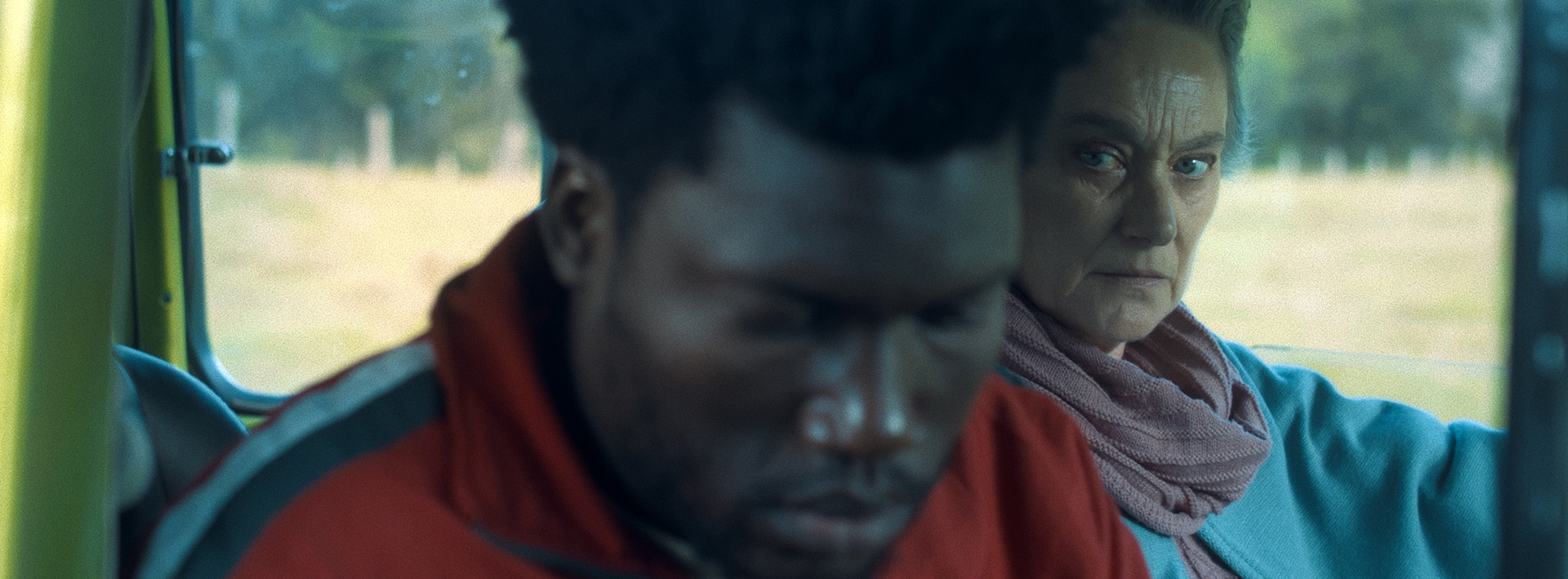 Em cena do filme, Porto Príncipe, estão dentro de um carro um homem negro de casaco vermelho e uma mulher branca, grisalha, de caso azul e echarpe rosa claro.