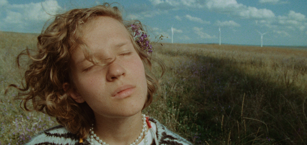 Uma jovem está no campo, com flores no cabelo. Ela está de olhos fechados, sentindo o vento.