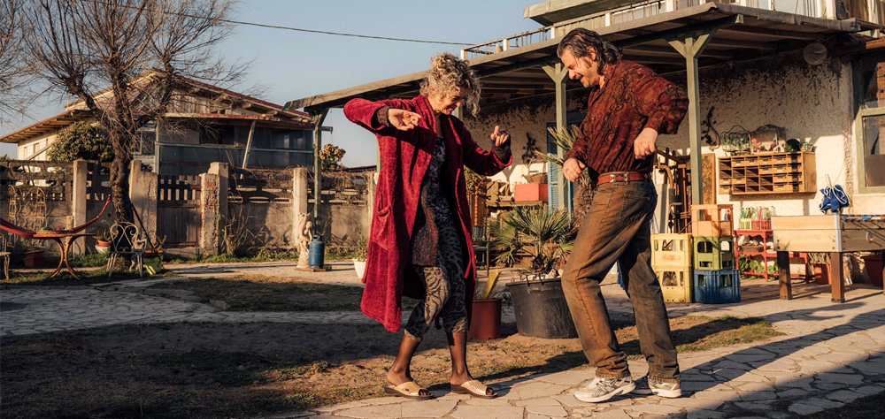 Em frente a uma casa, um homem e uma mulher dançam. Ele usa camisa e calça marrons, e ela veste um casaco vermelho e comprido.