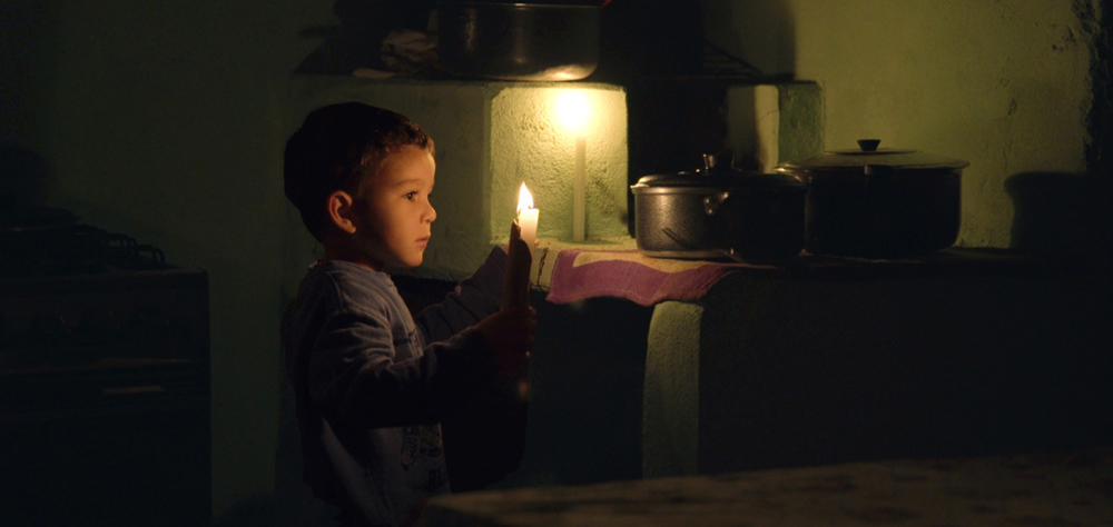 Um menino pequeno segura uma vela, em uma cozinha escura, e olha para panelas