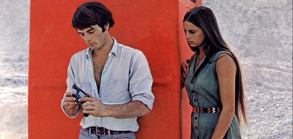 Cena de Zabriskie Point mostra um homem de camisa branca, mexendo em revólver. Uma mulher, de cabelos compridos, está ao seu lado e o observa.