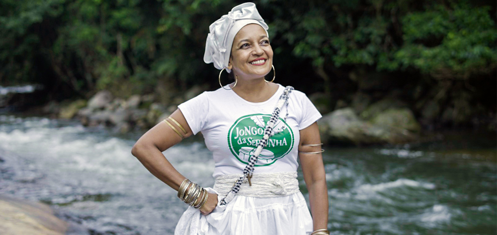 Cena de Razões Africanas mostra uma mulher, parada em frente a um rio. Ela usa um turbante branco e veste camiseta e saia brancas.