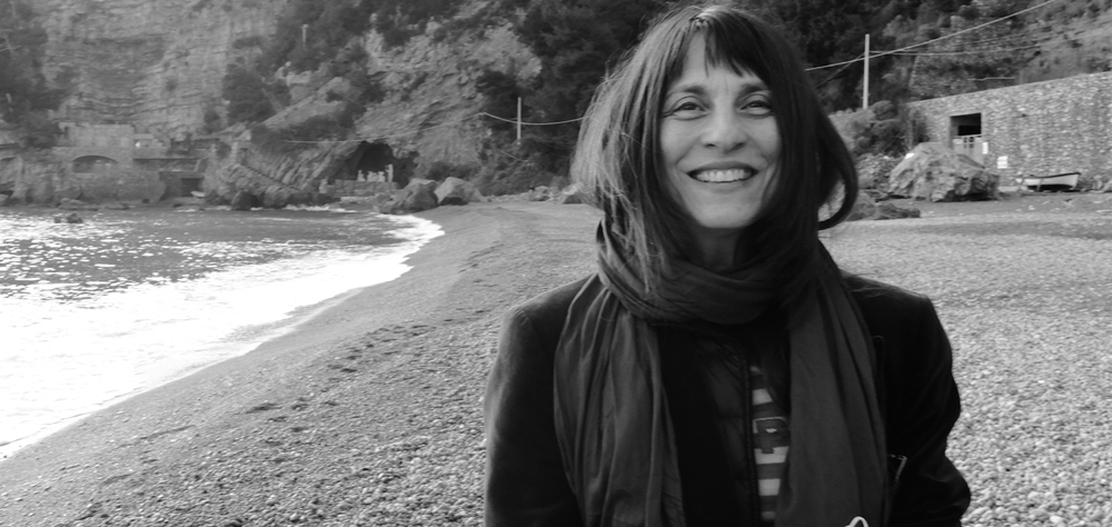 Foto preto e branca de Pascale Ramonda, em uma praia. Ela está sorrindo e veste cachecol e casaco.